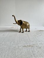 Front side view of a vintage brass elephant figurine with trunk up and etched decorative patterns on its body, placed on a textured white surface against a plain wall.