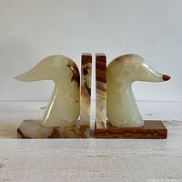 Side view of two carved stone bird head bookends, light cream with brown accents, positioned facing each other showing profile of bird heads and bases.