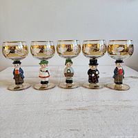Front view of all five vintage Goebel wine glasses with figurine bases and gold trimmed bowls etched with grapevine designs.