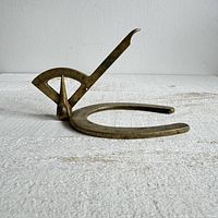 Side view of the vintage brass horse shoeing gauge showing the horseshoe base and the adjustable measuring arm with engraved graduations and pivot point.