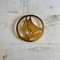 Top-down photo showing the circular solid brass trivet with a sailboat cut-out design resting on a wooden surface.