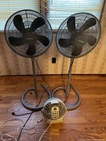 Two black-bladed standing Holmes floor fans and one small white portable fan on wooden floor in front of a window with blinds and floral wallpaper.