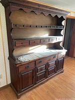 Full view of hardwood two-piece hutch showing upper shelving with scalloped edges and lower cabinets with ornate metal handles. Piece is positioned against a wall with wood flooring.