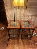 Two matching wooden end tables with glass insets on top and bottom shelves. One table has a black attached floor lamp missing its shade. Surfaces show light wear and scratches.