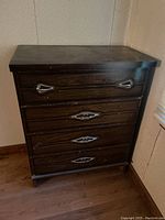 Front view of the small wood dresser showing four drawers with decorative metal handles, positioned against a wall in a corner.