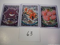 Three Pokemon cards next to each other: Gengar #94 on left, Kingler #99 center, Exeggcute #102 right, placed on white background with number card '63' in front.
