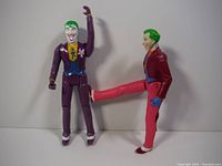 Two Joker action figures standing side by side, showing detailed paint and costumes in purple and red suits.