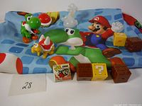 Photo of vintage Super Mario toys and figures on Mario-themed cloth, includes Yoshi, Koopa Troopa figures, and block pieces.