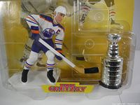 Close-up of Wayne Gretzky figure in Edmonton Oilers uniform holding hockey stick with Stanley Cup trophy beside it inside original plastic packaging.