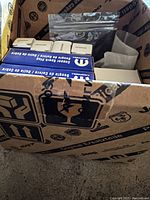 Open cardboard box containing several Mopar branded blue and white spark plug boxes labeled 'Copper Spark Plug', along with plastic bags and parts for installation.