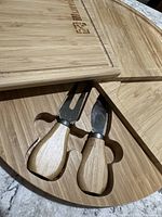 Close-up photo of the slide-out storage compartment open on the bamboo cutting board, showing two serving utensils with wooden handles — one fork and one knife, both designed for cheese.