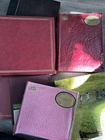 Top view of multiple photo albums including red leather-like, maroon, pink Designer Collection with sticker, and album with children playing cover.