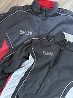 Two KooGa rugby zip-up jackets laid flat, one dark gray and red XXL and one black Large with white piping, showing front and collar detail.