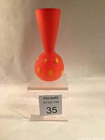 Front view of the satin orange-red glass vase with yellow polkadot design on a clear stand showing size and shape.