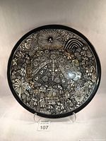 Full view of round black and white hand-painted narrative platter showing detailed figures, animals, and celestial motifs.