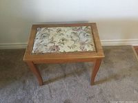 Front view of wooden bench with floral tapestry upholstered top and wooden curved legs on carpeted floor.