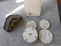 All 4 ceramic coasters with raised design, bottom of one coaster showing manufacturer sticker, original box with some discoloration, blue ceramic napkin holder with floral design.