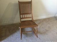 Front view of wooden rocking chair with wicker seat and backrest on carpeted floor against a plain wall.
