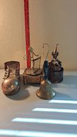 Four brass musical collectibles including a shoe music box, tennis player seated on chair music box, clown music box, and a bell placed on a white surface with a ruler in the background for scale.