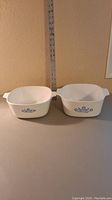 Two white Corning Ware cookware dishes side by side with blue cornflower design and measuring tape behind them.
