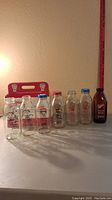 Photo showing six clear and one amber vintage milk bottles and a red cardboard milk bottle holder behind them on a flat surface with a measuring tape for scale.