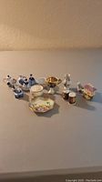 Miniature porcelain and ceramic items displayed on a flat surface, various figurines, bowls, cups, and other decorative pieces.