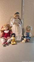 Four porcelain dolls including an angel doll standing with wings, a red dress doll seated, a small boy doll in blue overalls seated, and a small dog figure, all in front of a ruler for size reference.