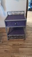 Front view of side table showcasing metal frame, purple wicker drawer with knob, and lower wicker shelf.