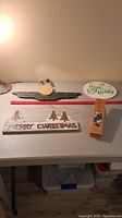 Four wooden decorative holiday-themed items arranged on a table against a wall. Includes a round 'Give Thanks' sign, angel decoration, Christmas tree with 'Merry Christmas' sign, and a large clip with painted flowers.