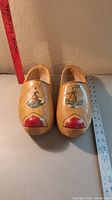 Top view of pair of wooden clogs showing painted windmill design on each toe and red tip accent, positioned next to rulers.