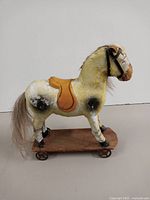 Side view showing yellow paper mache horse with black spots, orange saddle, horsehair mane and tail, and wooden cart base with wheels