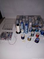 Clear plastic storage boxes with many spools of thread in a variety of colors arranged on a surface. Includes multiple small bobbins and large cones of thread.