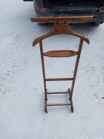 Front view of the medium brown wooden clothes hanger stand showing the broad hanger, central tray, vertical rods, horizontal base bar, and small wheels on snow ground.
