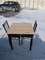 Photo showing kitchen table with light wood top and two black metal chairs on snowy outdoor ground.