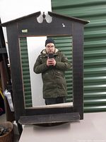 Front view of large rectangular mirror featuring a black wooden frame with a peaked top and small carved detail at the peak. Person taking photo visible in reflection.
