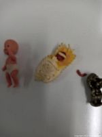Three miniature figures displayed on a white surface: one celluloid baby doll missing one arm and wearing a diaper; a yellow swaddled baby doll; and a small pewter troll figurine.
