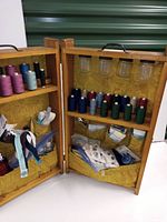 Open wooden sewing kit showing spools of thread, storage jars, and fabric pockets filled with sewing accessories.