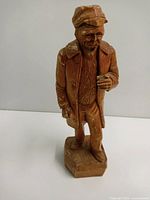 Front view of the signed wooden folk art statue showing the man's posture, hat, and facial features with some damage visible.