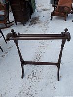 Wooden quilt/blanket rack standing on a snowy ground with minor scrapes visible on surface, showing front side with two horizontal bars and decorative finial tops.