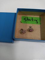Top view of two small clip-on sterling silver earrings with pink stones inside a blue box labeled 'Sterling'.