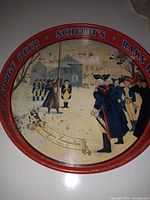 Photo of round metal beer tray with red rim and historical scene center showing military figures in Colonial-era uniforms and buildings in background; text on rim advertising Valley Forge Beer, Scheidt's, and Rams Head.