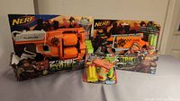 Photo showing two boxed Nerf Zombie Strike blasters side by side with small foam dart blaster toy in front.