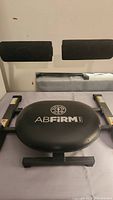 Front view of the AB Firm abdominal exercise machine showing the padded seat with logo and both arm supports with padding on a grey frame.