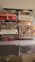 Box tops of stacked used puzzles showing multiple themes and brands including Hershey's, Ann Stookey, and Dowdle