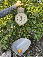 Photo of antique hanging grocery store scale with dial, metal chains and metal basket containing an orange on the basket.