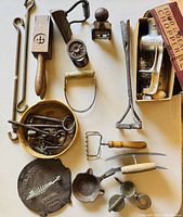 Group photo of assorted antique kitchen gadgets and tools spread on flat surface showing bacon press, chopper, keys, basket with nails, juicer, date stamp, and more