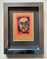 Framed Styrofoam print with black face image on rust-colored background in silver metal frame.