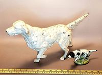 Two antique painted metal hunting dog figurines, large white dog with chipped paint and smaller black and white dog on green base, ruler for size reference.