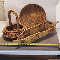 Three baskets displayed including a circular coiled basket with multicolor rings, a large wicker basket with handle, and a canoe-shaped gambella basket with brown and natural fiber patterns