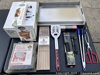 Overview photo showing boxed cedar grilling planks, boxed wooden skewers, metal skewers, metal spatula, two pairs of tongs, grill cleaning brushes, and folded BBQ grill cover.